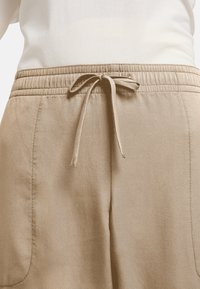 Pantalon beige avec une taille élastique et un cordon de serrage. Comprend des poches latérales et une texture lisse, photographié en vue arrière.