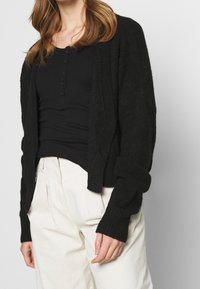 Svart, texturerad cardigan med en lös passform och sänkta axlar. Kombinerad med en svart åtsittande topp och vita högmidjade byxor.