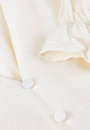 Close-up van crèmekleurige stof met twee bedekte knopen en een geplooid elastiekdetail in de rechterbovenhoek.