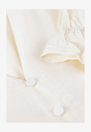 Close-up van crèmekleurige stof met twee bedekte knopen en een geplooid elastiekdetail in de rechterbovenhoek.
