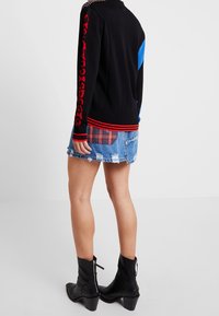 En person som bär en svart tröja med röd text på ärmen, en slitna denimminikjol med en rutmönstrad ficka och svarta klackstövletter.