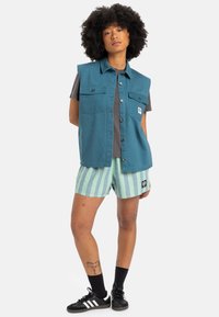 Teal ärmlös väst med två bröstfickor, grå t-shirt under, och gröna randiga shorts. Svarta sneakers med vita ränder.