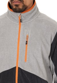 Uomo che indossa una giacca zip-up grigia e nera con zip arancione e dettagli sulla tasca del petto, mostrando la parte bassa del viso e il collo.