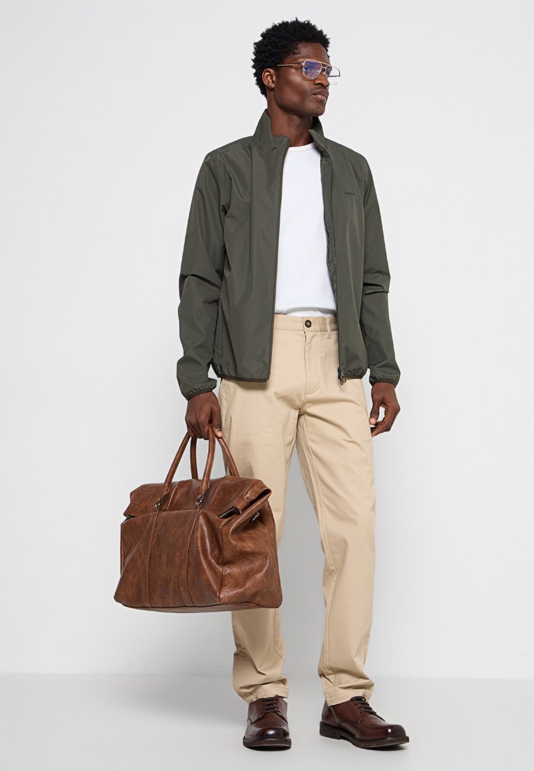 Giacca verde oliva, t-shirt bianca, pantaloni beige, borsa a tracolla in pelle marrone, scarpe marroni e occhiali rettangolari. Abbigliamento moderno e casual.