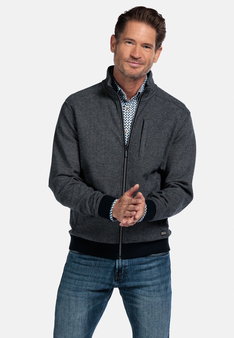 Graue Zip-Jacke mit strukturierter Oberfläche, schwarzen gerippten Bündchen und Saum sowie einer Brusttasche. Getragen über einem blauen gemusterten Hemd.