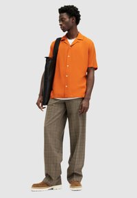 Camisa naranja de manga corta con botones, combinada con pantalones cortos de cuadros marrones de pierna ancha. El hombre lleva una bolsa tote negra y usa zapatos marrones con suela gruesa.