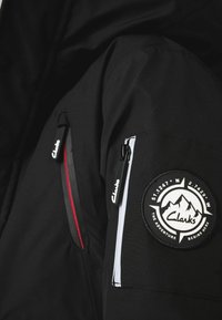 Veste noire avec fermeture éclair rouge, poche latérale et un patch circulaire brodé représentant des montagnes et du texte. Le matériau semble lisse et durable.