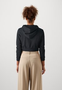 Persona con cabello rizado, vistiendo una sudadera corta negra y pantalones de tiro alto beige de pierna ancha, de pie de espaldas contra un fondo blanco.