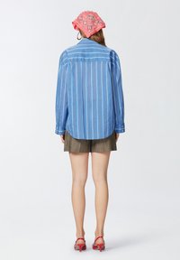 Camicia a maniche lunghe a strisce blu chiaro con polsini a bottone, abbinata a pantaloni corti marroni a pieghe e sandali con tacco rossi; sciarpa rosa a motivo in testa.