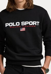Sort sweatshirt med "POLO SPORT" og "RALPH LAUREN" i hvid og rød tekst. Har et lille amerikansk flagdetalje. Materialet er en blanding af bomuld.