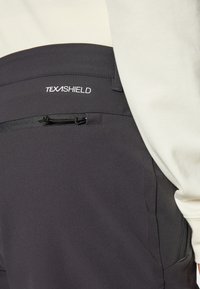 Close-up van een zwarte broekzak met rits en TEXASHIELD-logo, gedragen met een lichtkleurig overhemd met lange mouwen.