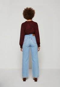 Kvinna med lockigt hår står vänd mot en ljusgrå vägg, iförd en mörkröd blommig blus, ljusblå högmidjade jeans och bruna klackskor.