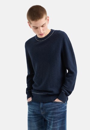Maglione testurizzato blu navy con design a nido d'ape, collo girocollo e cuciture bianche a contrasto nel colletto e nei polsini. Abbinato a jeans.