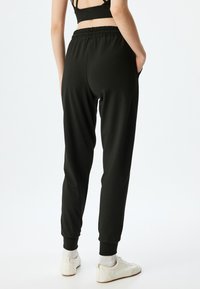 Zwarte joggers met elastische tailleband en taps toelopende boorden, voorzien van zijzakken, gemaakt van een gladde stof. Gedragen met witte sneakers.