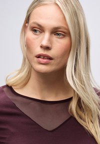 Blusa con un profondo scollo a V e inserto in rete trasparente al colletto. Il colore è un ricco bordeaux, con una texture liscia e un design aderente.