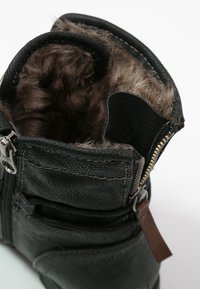 Botte en cuir noir avec doublure en fourrure marron visible, partiellement déboutonnée, montrant une fermeture éclair en métal et une languette en cuir.