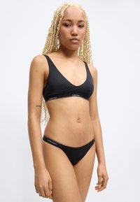 Γυναίκα με μακριά ξανθά πλεξούδες που φοράει μαύρο σουτιέν Calvin Klein και ταιριαστό εσώρουχο, στέκεται μπροστά σε απλό φωτεινό φόντο.