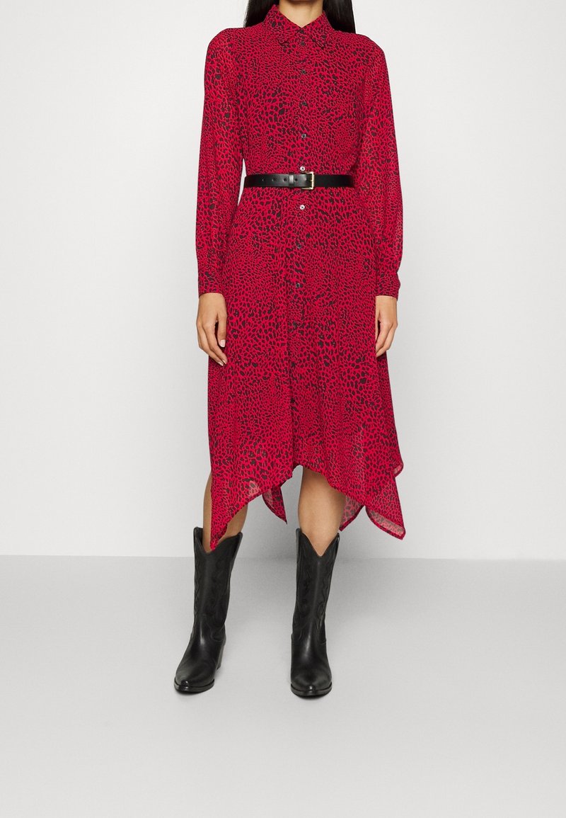 Femme portant une robe midi rouge à imprimé léopard avec un ourlet asymétrique, une ceinture noire et des bottes de cowboy noires, debout devant un fond uni.