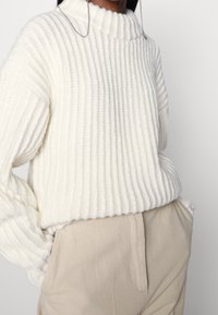 Personne portant un pull en maille côtelée crème avec un décolleté festonné et un pantalon beige taille haute, les mains dans les poches.