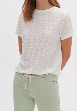 Vrouw draagt een witte, gestructureerde top met korte mouwen en lichtgroene broek met een witte touwriem in een strik aan de taille.
