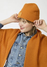 Orange knit beanie with ribbed texture and a square logo patch, paired with an open-knit orange wrap and a denim jacket.