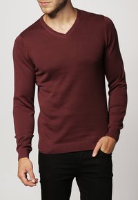 Bordeauxrode v-hals trui met lange mouwen, gemaakt van een zachte gebreide stof. Heeft geribbelde manchetten en zoom, die een aansluitende pasvorm creëren.