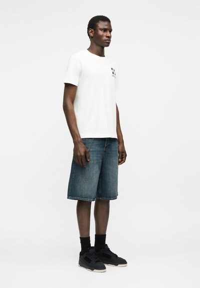 Άνδρας που στέκεται φορώντας λευκό κοντομάνικο μπλουζάκι, τζιν σορτς μέχρι το γόνατο, μαύρες κάλτσες και μαύρα αθλητικά παπούτσια σε λευκό φόντο.