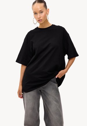 Femme avec des cheveux coiffés en arrière, portant de grandes boucles d'oreilles en anneau, un t-shirt noir surdimensionné et un jean gris ample, se tenant avec une main dans la poche.