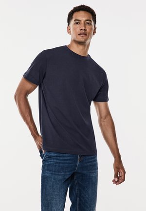 Dunkelblaues T-Shirt aus Baumwolle mit Rundhalsausschnitt und kurzen Ärmeln, kombiniert mit blauen Jeans. Glatte Textur, lässige Passform.