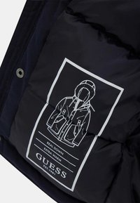 Juodas pūkinis švarkas, viduje esantis baltas etiketas su grafiniu paltu, tekstu "TIKRAS PUŠTAS" ir "PAPILDOMAI ŠILTAS", bei prekės ženklu "GUESS."