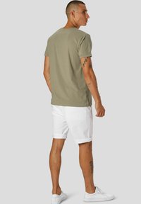 Oliven grønn t-skjorte med klassisk passform, kombinert med hvite shorts. Modellen har synlige tatoveringer på armene og benet, og har på seg hvite sneakers.