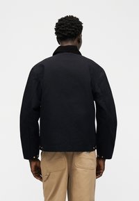 Moški, ki nosi črno jakno z zapetimi manšetami in bež hlače z velikimi zadnjimi žepi, stoji pred enobarvnim belim ozadjem.