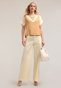 Top senza maniche color tan con scollatura a V, maniche corte color crema, pantaloni larghi color crema, sandali gialli e una borsa bianca con dettagli neri.