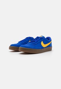 Ténis de camurça azul com detalhes amarelos, atacadores planos e sola de borracha castanha. Bico arredondado e textura suave, com o logo swoosh.