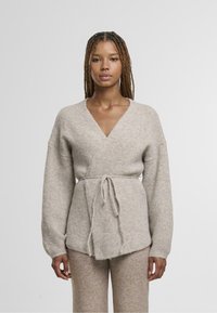 Smėlio spalvos megztinis apsiaustas su gilia V formos iškirpte, ilgomis rankovėmis ir diržu liemens srityje. Minkšta tekstūra su švelniu pūkeliu.