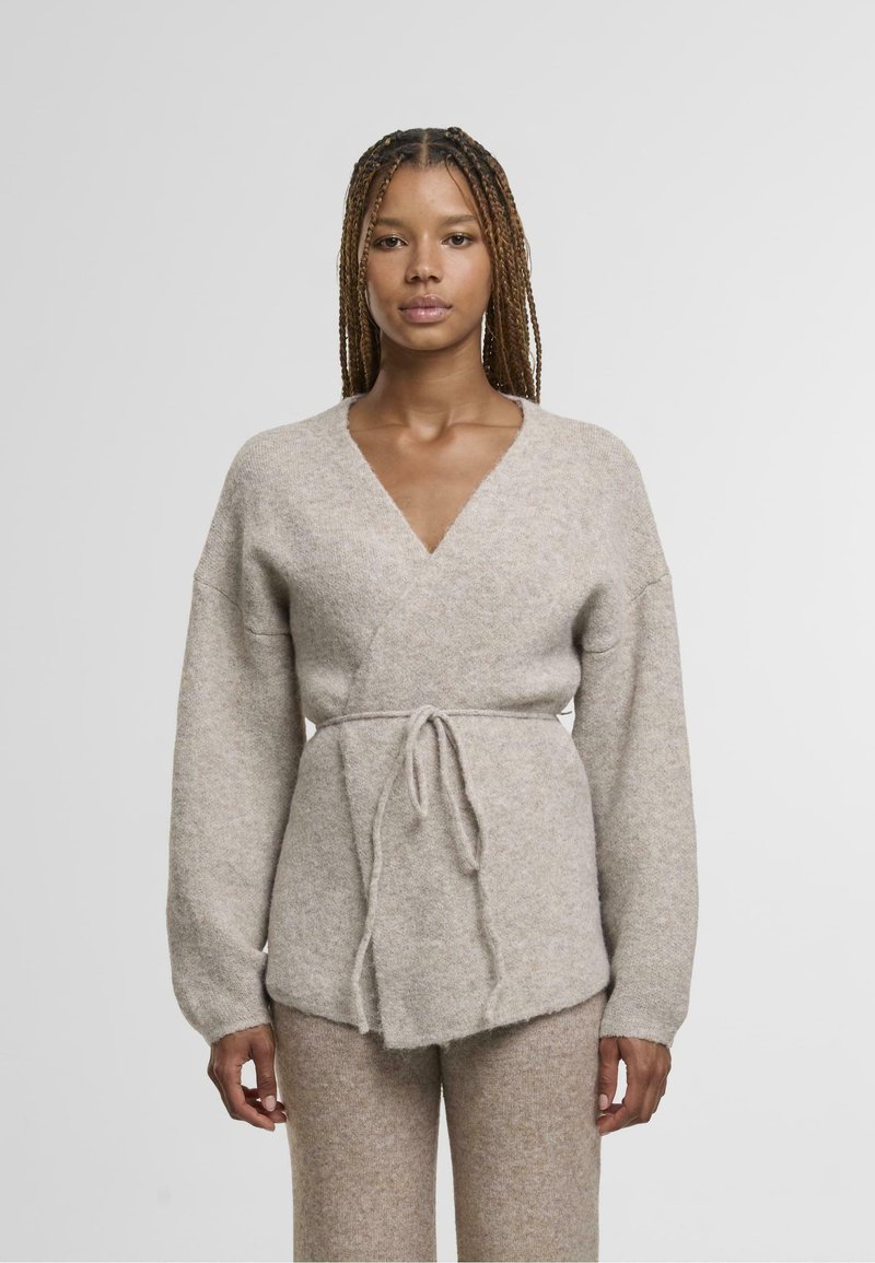 Beżowy sweter z dzianiny, z głębokim dekoltem w szpic, długimi rękawami i paskiem wiązanym w talii. Miękka struktura o lekko puszystym wykończeniu.