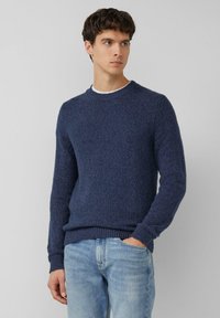 Granatowy sweter z dzianiny o fakturowanym wykończeniu, z okrągłym dekoltem i prążkowanymi mankietami. Połączony z jasnoniebieskimi dżinsami, stoi na neutralnym tle.