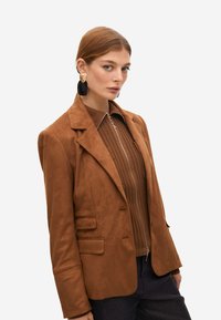 Blazer de ante marrón con un ajuste entallado, que presenta solapas de muesca, dos bolsillos frontales con solapa y una textura suave. El modelo lleva una camiseta marrón de canalé debajo.