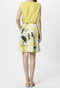 Gul bluse med samlede halsudskæring og snore; parret med en blomsterprint nederdel i gule, sorte og hvide farver; flade beige sandaler.