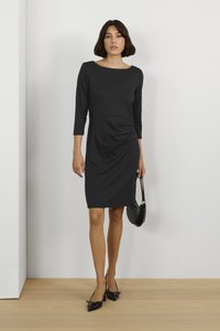 Abito nero al ginocchio con maniche a tre quarti, scollatura rotonda e dettagli raccolti sui lati. La modella tiene in mano una piccola borsa nera e indossa scarpe nere a punta.