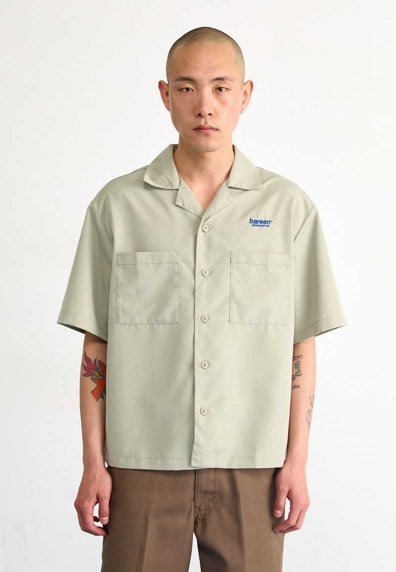 Camicia a maniche corte verde chiaro con bottoni, dotata di due tasche sul petto e logo. Tessuto liscio, vestibilità comoda e colletto arrotondato.