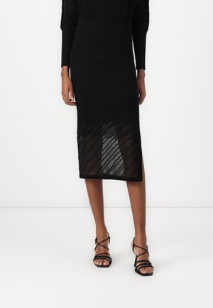 Femme portant une jupe midi noire texturée avec des panneaux transparents en diagonale et des sandales à talons noires à brides, debout sur un fond uni.