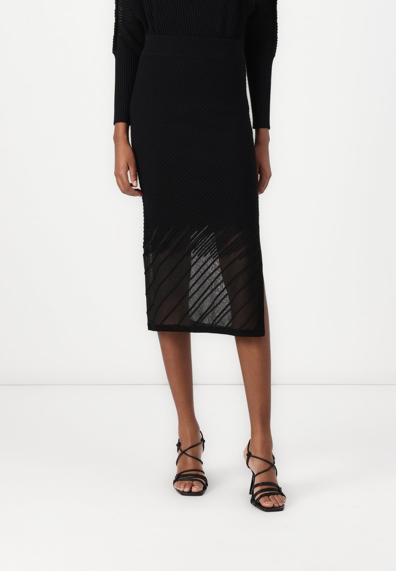 Femme portant une jupe midi noire texturée avec des panneaux transparents en diagonale et des sandales à talons noires à brides, debout sur un fond uni.