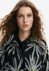 Floral-patterned black jacket with white and yellow daisies, textured fabric, and a zippered collar. Model has curly hair and a dark shirt.