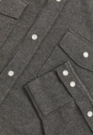 Tessuto a maglia grigio scuro con bottoni a pressione, tasche sul petto e un polsino con due bottoni a pressione, che dettaglia un primo piano di un indumento.