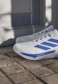 White running shoes with blue stripes and "DREAMSTRIKE+" text, placed on a stone pavement near a gray wall with shadows.