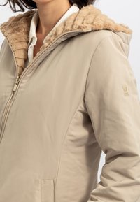 Giacca beige con zip e cappuccio in pelliccia sintetica testurizzata. Presenta un esterno liscio e un logo discreto sulla manica. Design semplice, senza motivi.