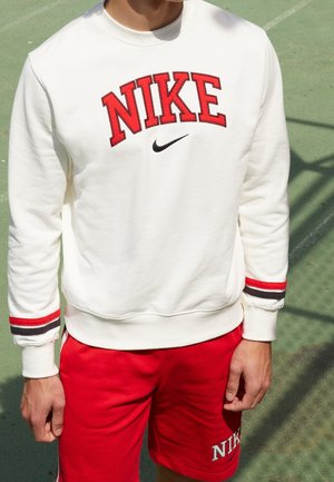 Osoba nosząca białą bluzę Nike z czerwonymi i czarnymi paskami na mankietach oraz czerwone spodenki Nike stoi na zewnętrznym boisku.