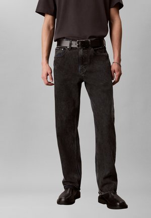 Hombre con vaqueros negros desgastados, cinturón negro, camiseta negra, pulsera plateada y zapatos negros, de pie contra un fondo gris.