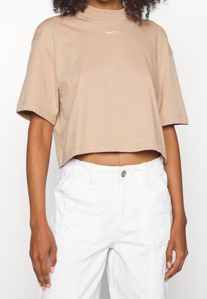 Donna che indossa un t-shirt Nike beige cropped e pantaloni bianchi con tasche frontali, in piedi contro uno sfondo neutro.